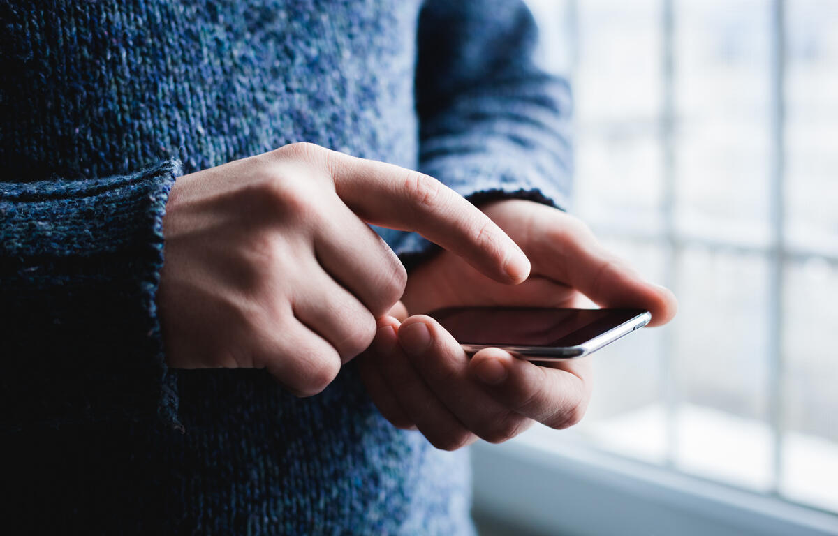 Hands of a person in a blue sweater using a smartphone indoors by a window.