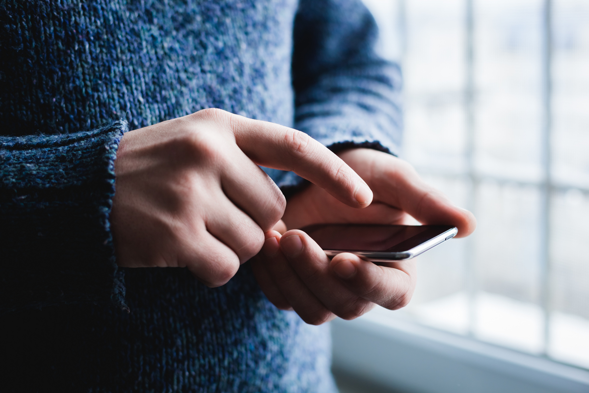 Hands of a person in a blue sweater using a smartphone indoors by a window.