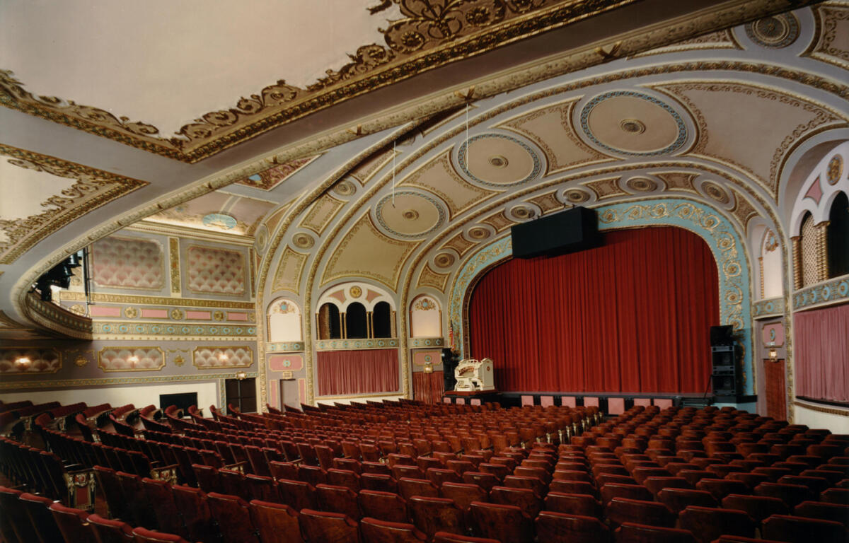 Historic ornate theater interior with red velvet seats facing a stage behind closed red curtains and gilded blue arches.