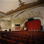 Historic ornate theater interior with red velvet seats facing a stage behind closed red curtains and gilded blue arches.