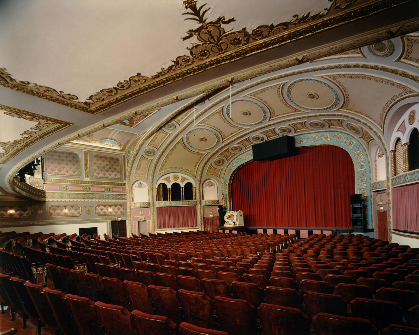 Historic ornate theater interior with red velvet seats facing a stage behind closed red curtains and gilded blue arches.
