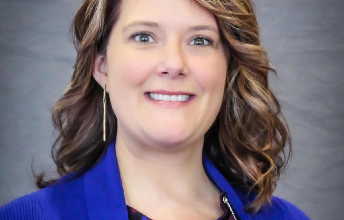 Smiling woman with wavy light brown hair wearing a royal blue blazer, photographed waist up against a gray backdrop.