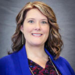 Smiling woman with wavy light brown hair wearing a royal blue blazer, photographed waist up against a gray backdrop.