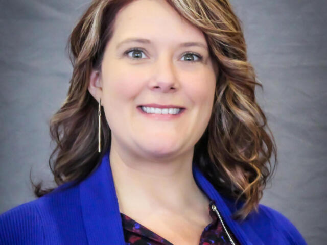 Smiling woman with wavy light brown hair wearing a royal blue blazer, photographed waist up against a gray backdrop.