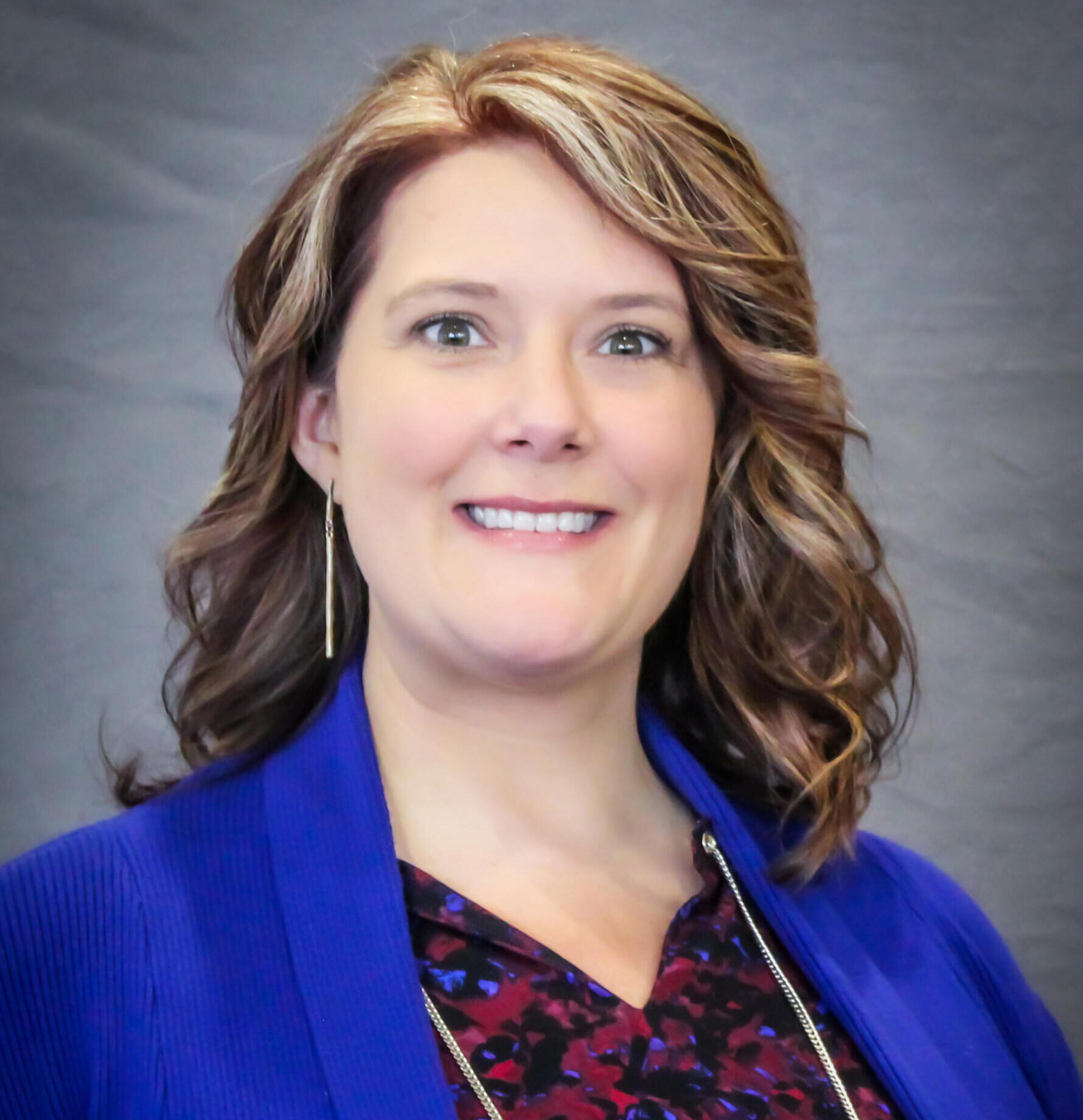 Smiling woman with wavy light brown hair wearing a royal blue blazer, photographed waist up against a gray backdrop.