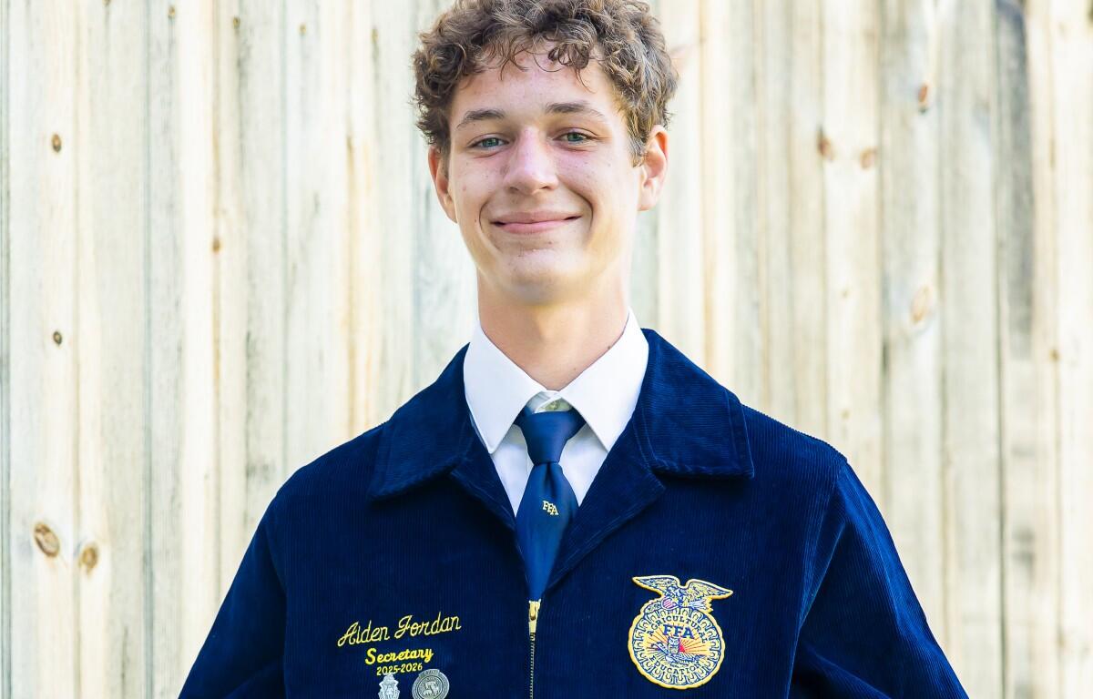 Young man in a navy FFA jacket smiling, posing for a portrait against a wooden fence; name reads Alden Jordan, Secretary 2025–2026, with FFA emblem nearby.