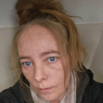 Close-up selfie of a woman with blue eyes, light skin, and a messy bun, wearing a dark jacket and scarf, looking at the camera.