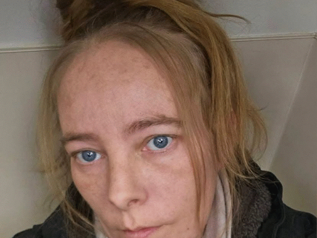 Close-up selfie of a woman with blue eyes, light skin, and a messy bun, wearing a dark jacket and scarf, looking at the camera.