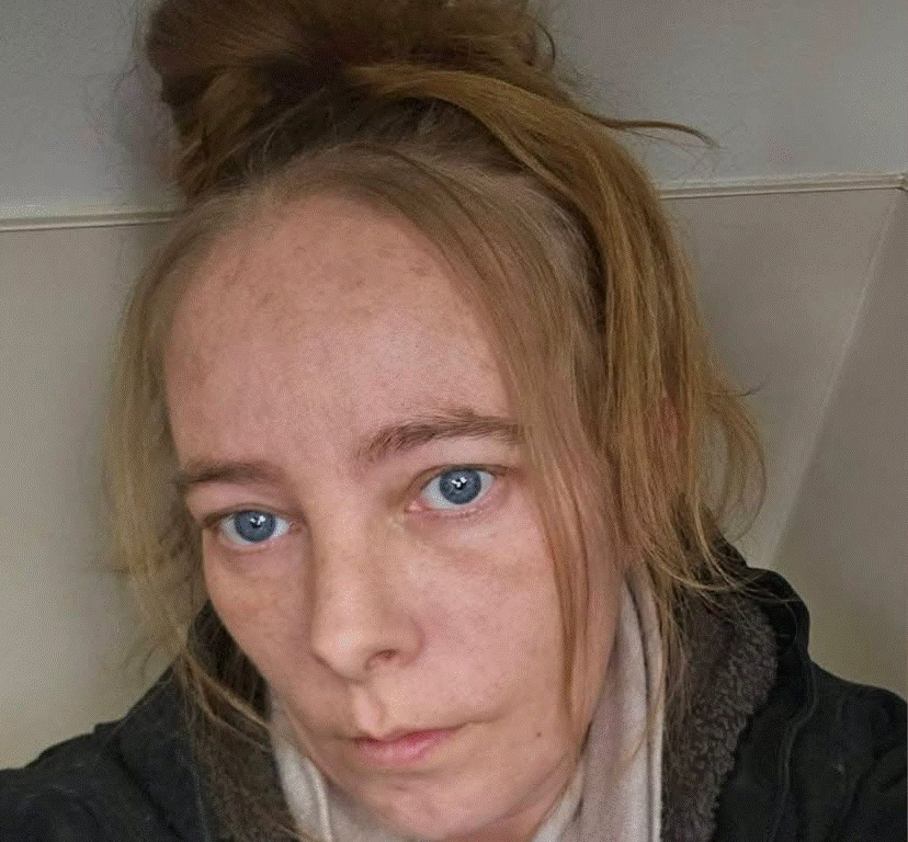Close-up selfie of a woman with blue eyes, light skin, and a messy bun, wearing a dark jacket and scarf, looking at the camera.