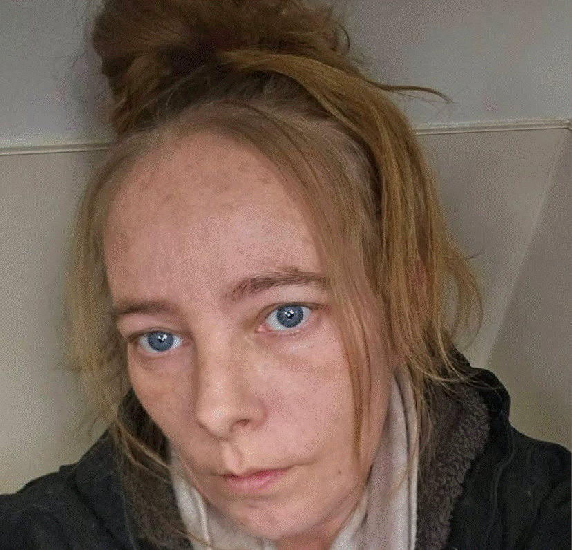 Close-up selfie of a woman with blue eyes, light skin, and a messy bun, wearing a dark jacket and scarf, looking at the camera.