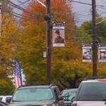 Street scene with banners featuring portraits hanging from utility poles amid autumn trees and cars on the road.