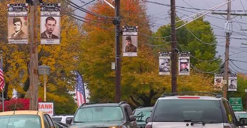 Street scene with banners featuring portraits hanging from utility poles amid autumn trees and cars on the road.