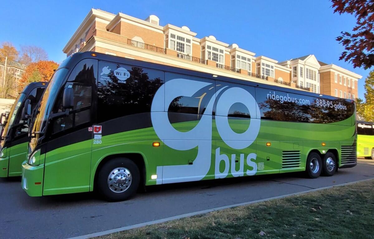 Green and black Go Bus charter bus with large 'go' lettering parked by a brick-campus building. Wi‑Fi sticker visible on window.