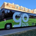 Green and black Go Bus charter bus with large 'go' lettering parked by a brick-campus building. Wi‑Fi sticker visible on window.