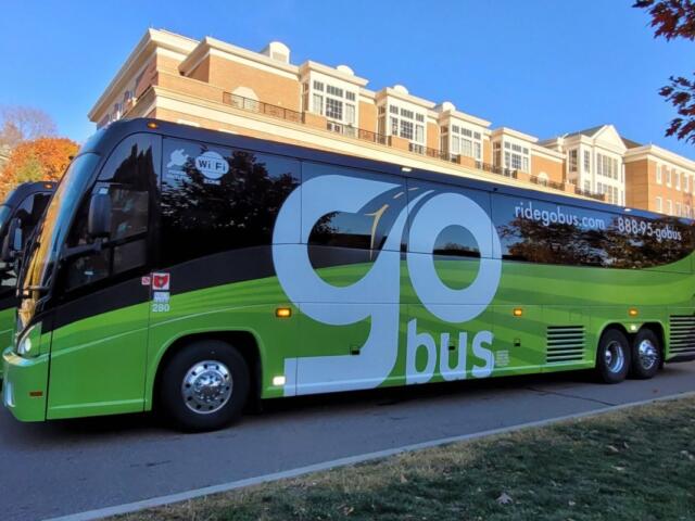 Green and black Go Bus charter bus with large 'go' lettering parked by a brick-campus building. Wi‑Fi sticker visible on window.