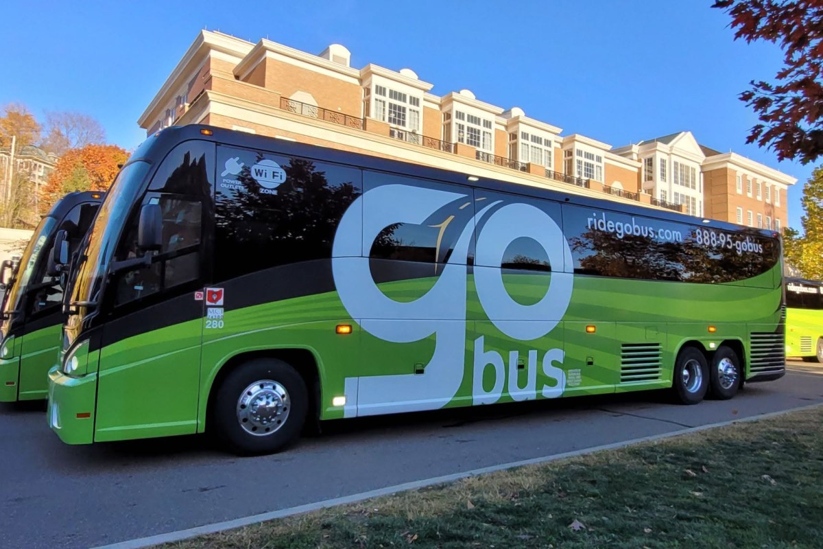 Green and black Go Bus charter bus with large 'go' lettering parked by a brick-campus building. Wi‑Fi sticker visible on window.
