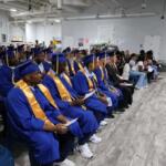 Row of graduates in blue gowns and caps with gold sashes seated in a classroom-style auditorium for a ceremony, holding programs as an audience watches.