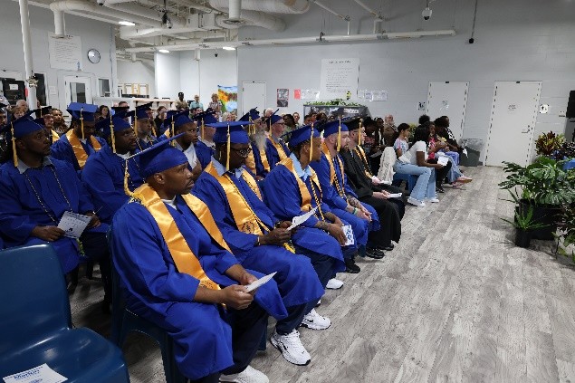 Row of graduates in blue gowns and caps with gold sashes seated in a classroom-style auditorium for a ceremony, holding programs as an audience watches.