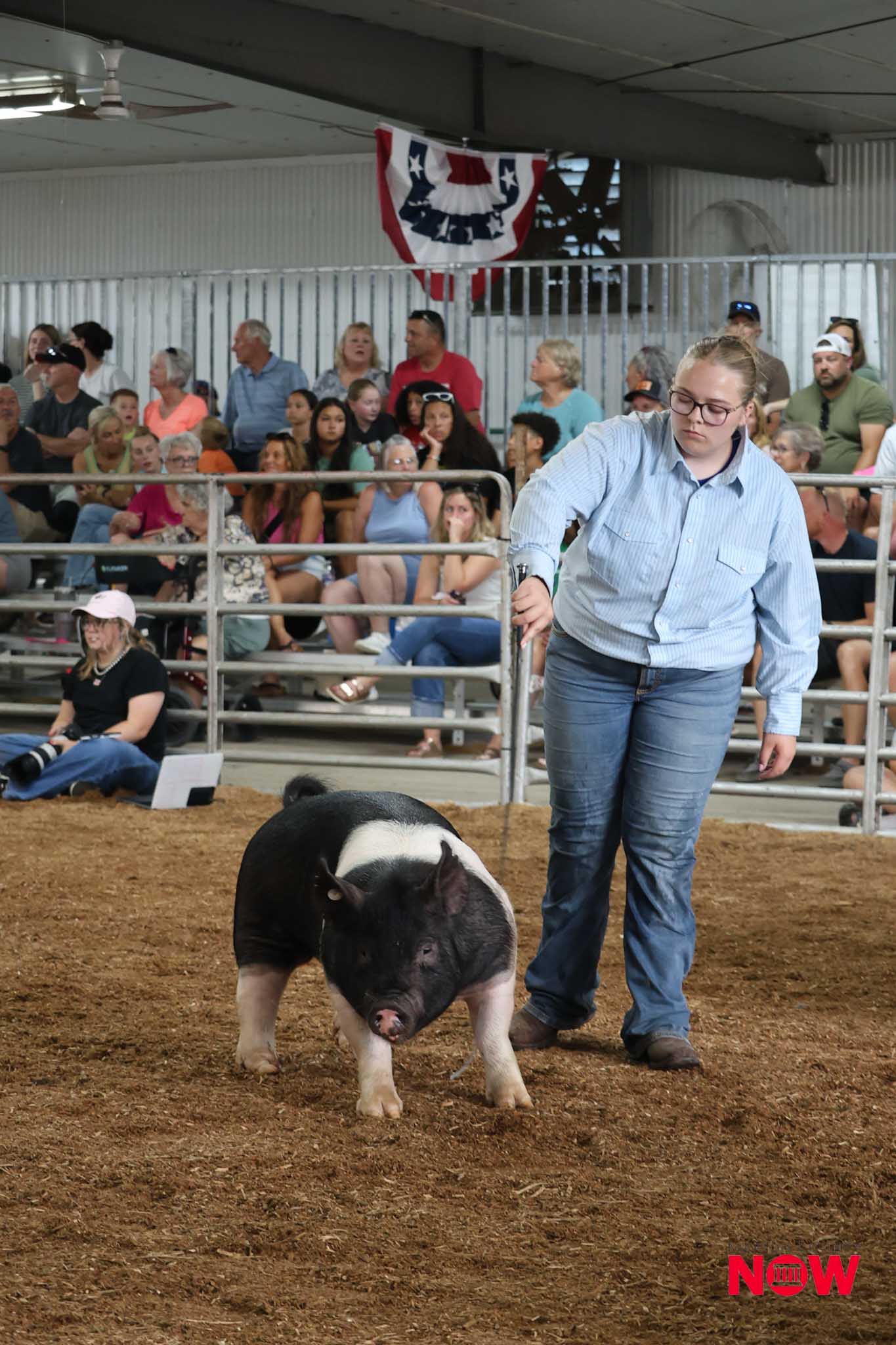 Silhouette of a person in a light blue shirt guiding a pig in a dirt arena as a large crowd watches from bleachers behind a metal railing, with a patriotic banner above.