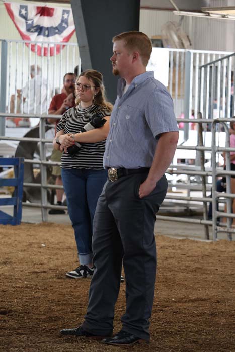 Man in a blue short-sleeve shirt and dark trousers stands with hands in pockets in a livestock arena.