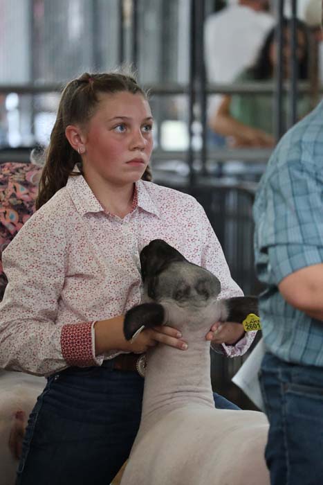 Young woman in a pink floral shirt holds a white lamb with a dark head at a livestock show.