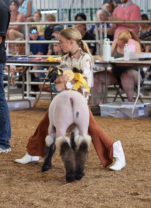 Young girl in western clothing kneels beside a small pony, holding a yellow ribbon at a fair with spectators in the background.