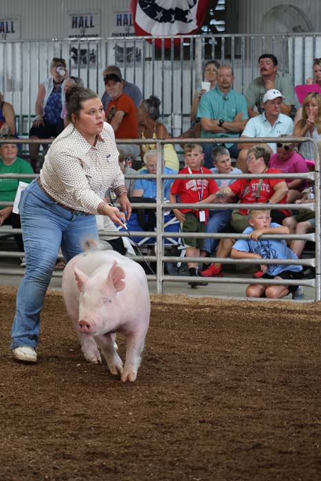 Young woman guiding a pink pig in a dirt arena at a livestock show, with spectators watching from the stands.