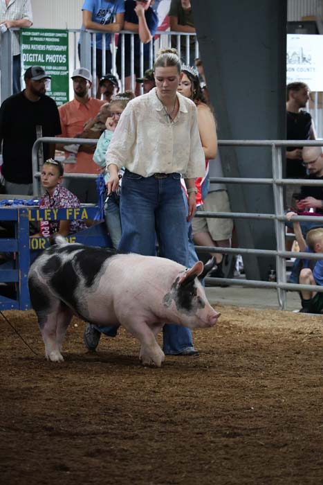 A woman guides a large black-and-white pig in a dirt arena, with spectators watching behind a metal railing.