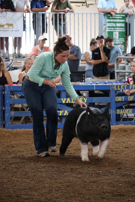 Woman in a mint-green top guides a black and white pig through a pen at a livestock show, with spectators watching in the background.