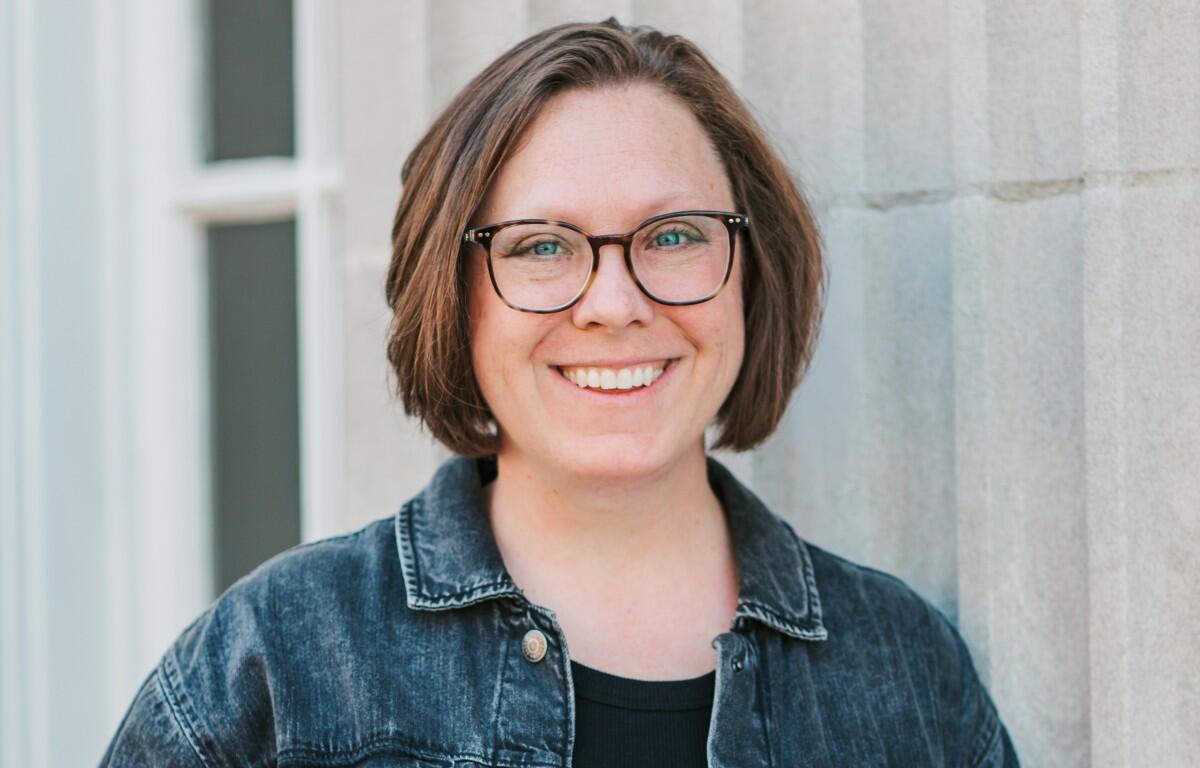 Smiling woman with short brown hair and black-framed glasses, wearing a denim jacket, against a light stone wall.