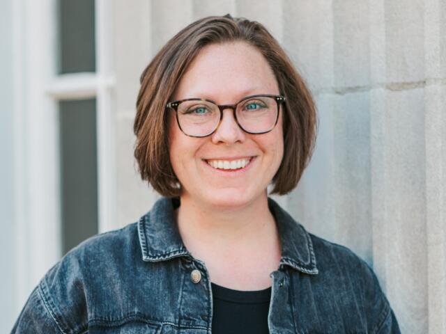 Smiling woman with short brown hair and black-framed glasses, wearing a denim jacket, against a light stone wall.