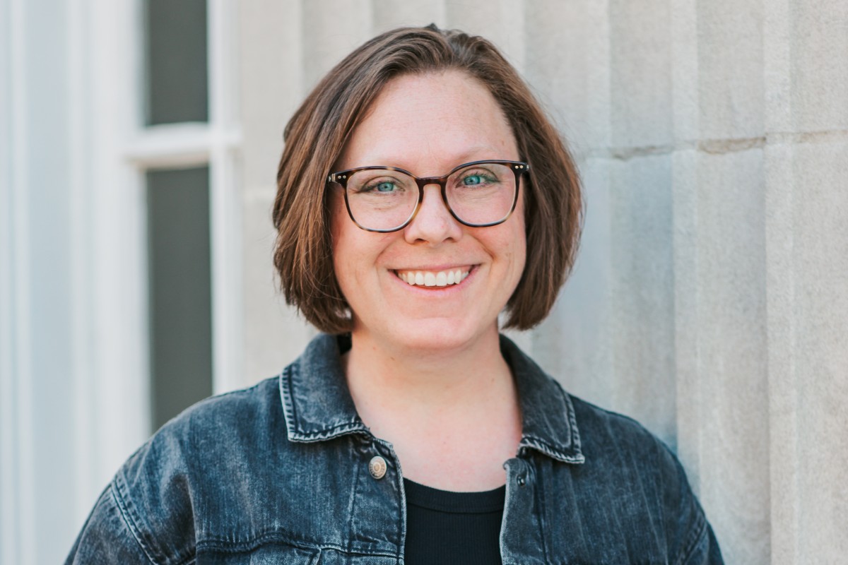 Smiling woman with short brown hair and black-framed glasses, wearing a denim jacket, against a light stone wall.