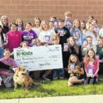 Large group of children posing outside a school building, holding snack-sized gift bags and a large ceremonial check for Kiwanis K-Kids, with a dog seated at the front.