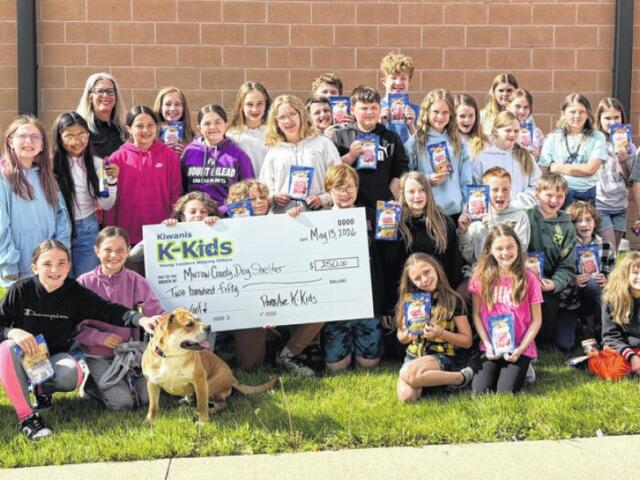 Large group of children posing outside a school building, holding snack-sized gift bags and a large ceremonial check for Kiwanis K-Kids, with a dog seated at the front.