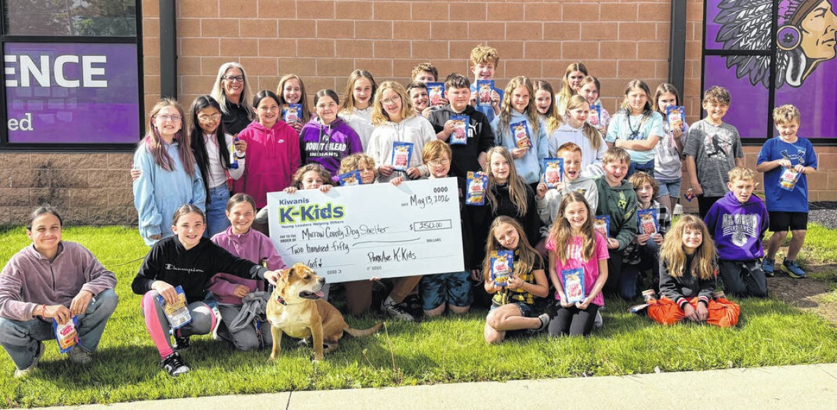 Large group of children posing outside a school building, holding snack-sized gift bags and a large ceremonial check for Kiwanis K-Kids, with a dog seated at the front.