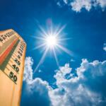 Wooden thermometer angled toward a bright sun in a clear blue sky with fluffy clouds, indicating hot weather