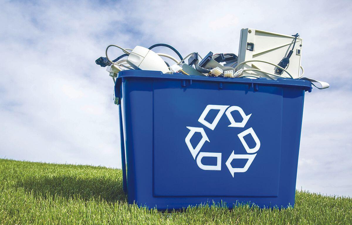 Blue recycling bin overflowing with discarded electronics outdoors on a grassy field under a blue sky.