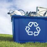 Blue recycling bin overflowing with discarded electronics outdoors on a grassy field under a blue sky.
