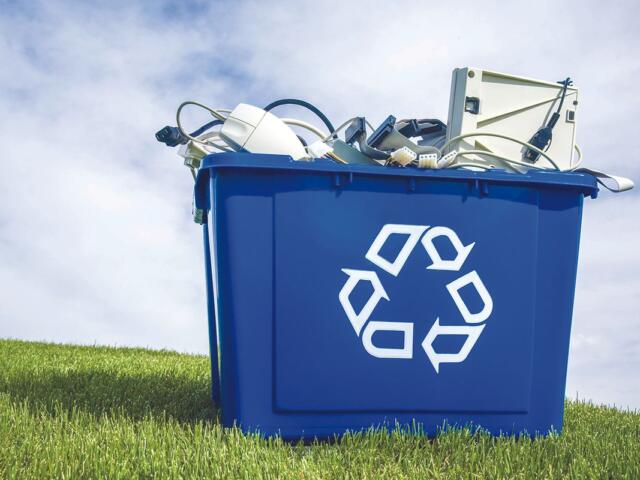 Blue recycling bin overflowing with discarded electronics outdoors on a grassy field under a blue sky.