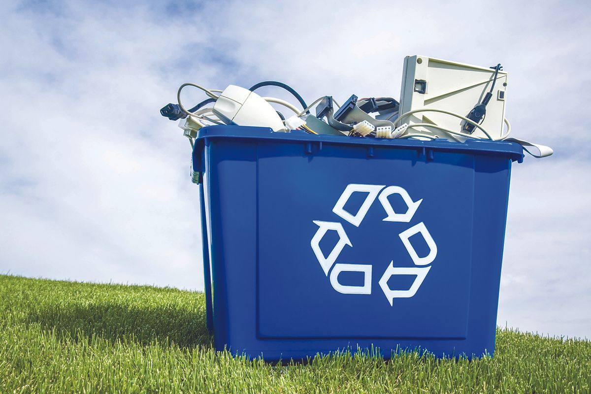 Blue recycling bin overflowing with discarded electronics outdoors on a grassy field under a blue sky.
