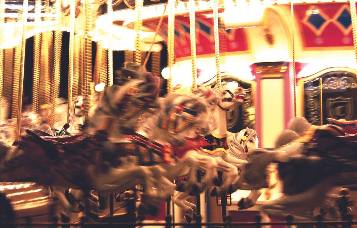 Blurred carousel in motion with white and painted horses, bright lights, and an ornate red-gold backdrop at night.
