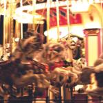 Blurred carousel in motion with white and painted horses, bright lights, and an ornate red-gold backdrop at night.