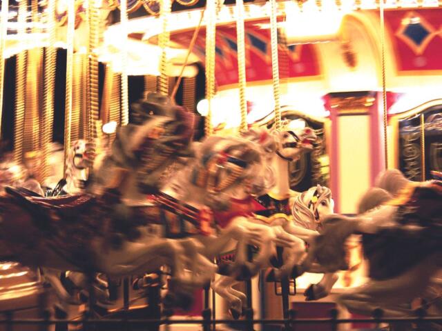 Blurred carousel in motion with white and painted horses, bright lights, and an ornate red-gold backdrop at night.