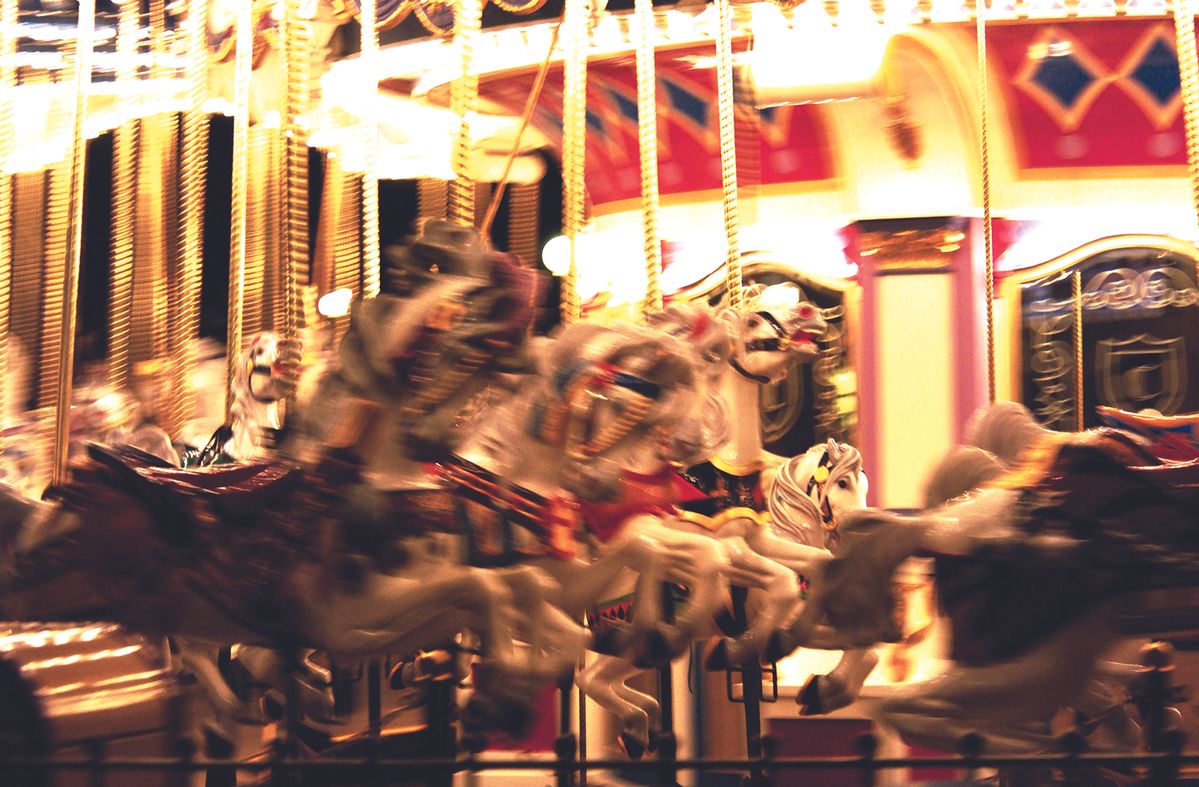 Blurred carousel in motion with white and painted horses, bright lights, and an ornate red-gold backdrop at night.