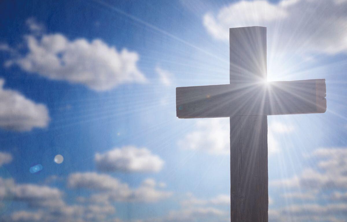 Cross silhouetted against a bright sky with sun rays streaming around it, symbolizing faith.