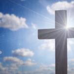 Cross silhouetted against a bright sky with sun rays streaming around it, symbolizing faith.
