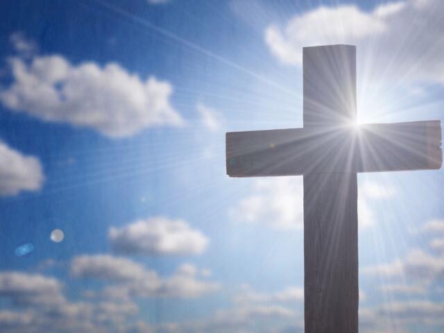 Cross silhouetted against a bright sky with sun rays streaming around it, symbolizing faith.