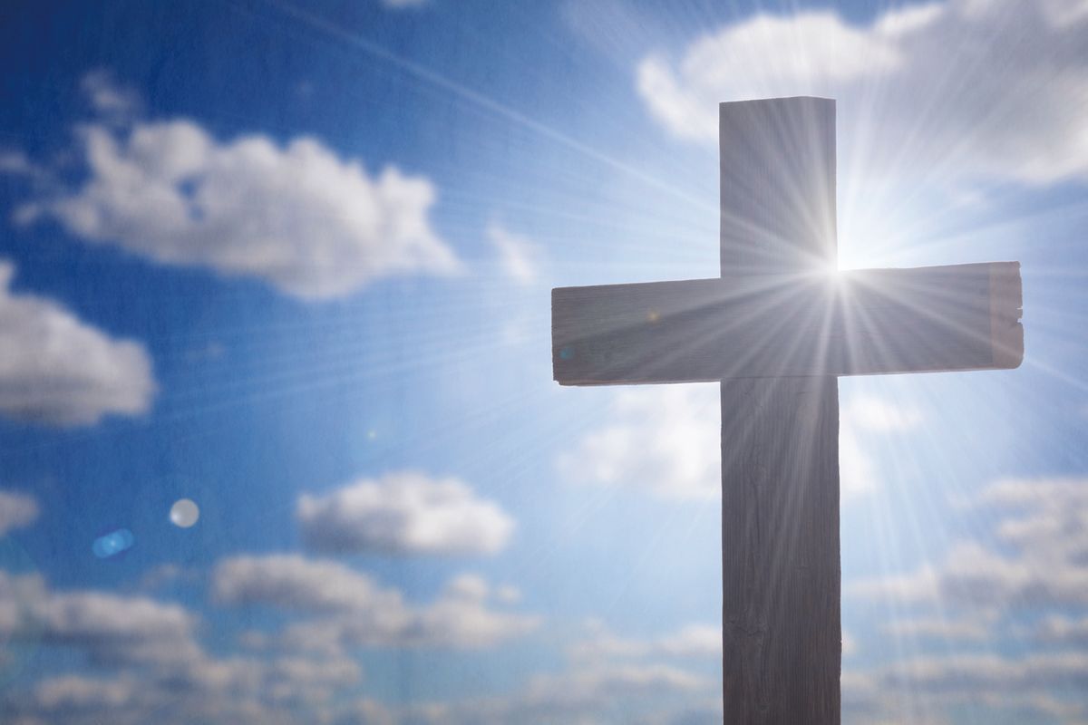 Cross silhouetted against a bright sky with sun rays streaming around it, symbolizing faith.
