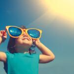 Young girl wearing bright orange oversized sunglasses, looking up at the sunny sky.