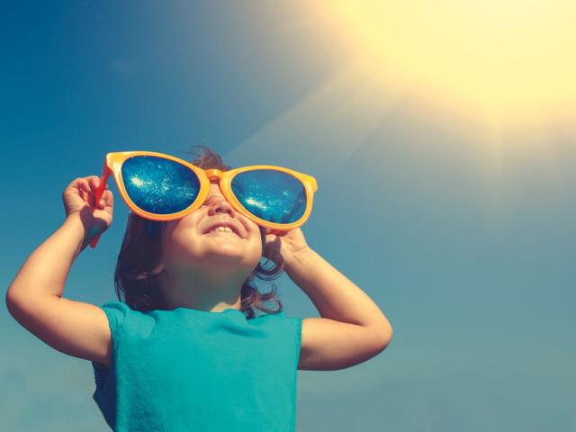 Young girl wearing bright orange oversized sunglasses, looking up at the sunny sky.